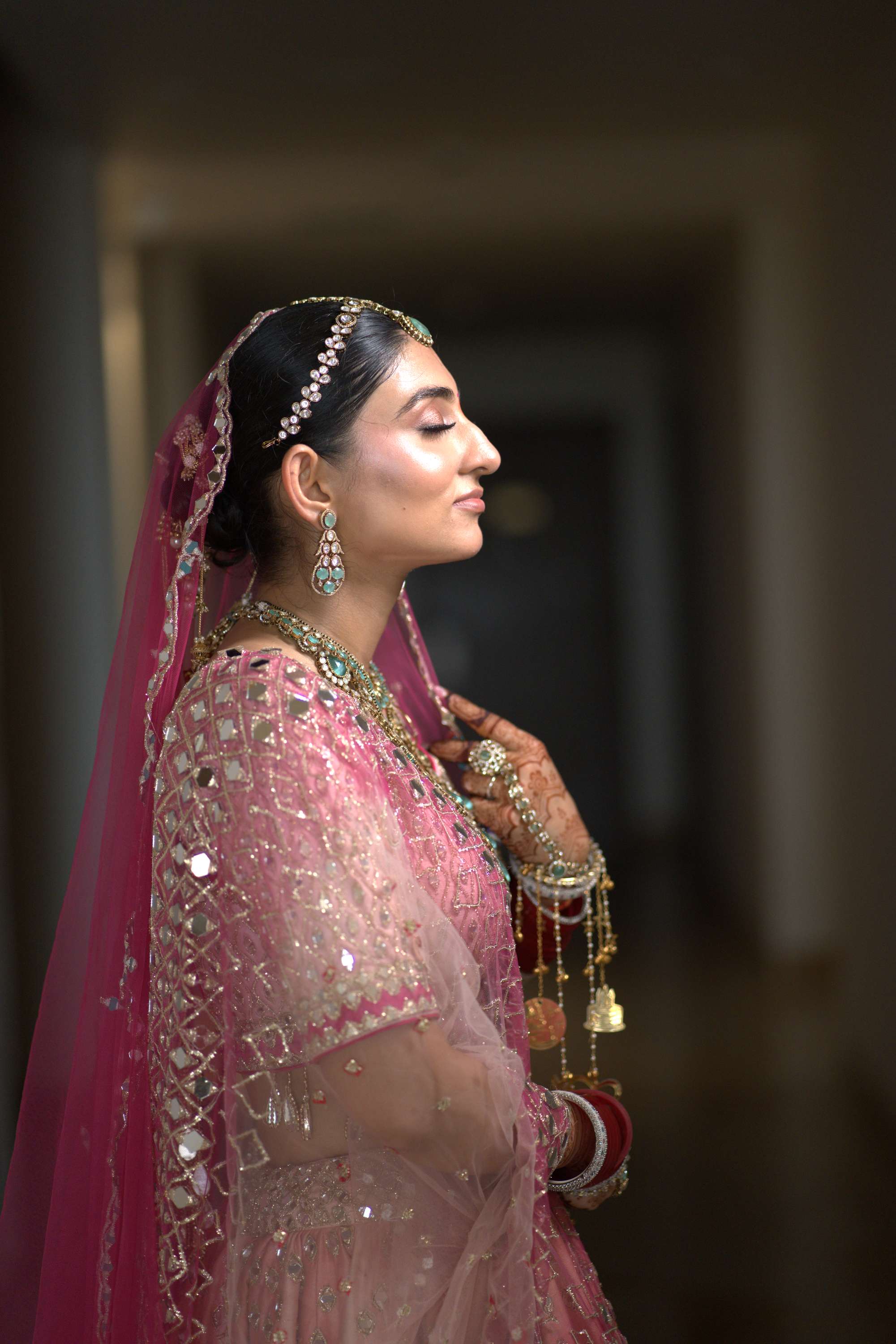 The bride in a pink lehenga, side profile in the dark hall